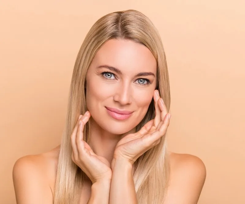A blonde woman with clear skin gently touching her face against a peach-colored background. - Acne
