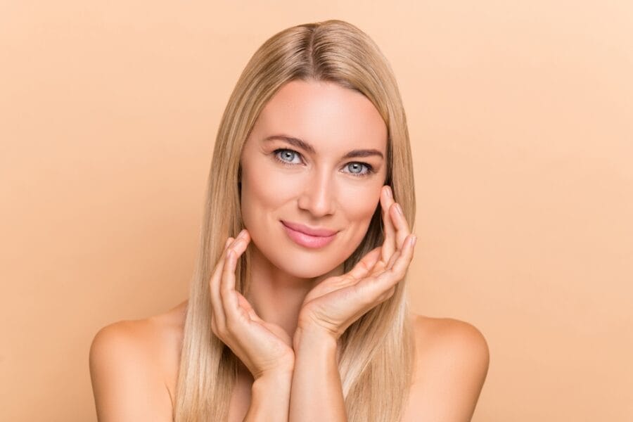 A blonde woman with clear skin gently touching her face against a peach-colored background. - Acne